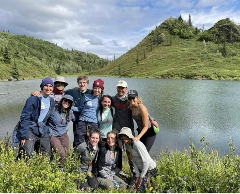 Group photo of teens in Alaska with green mountains and a blue swimming hole behind them