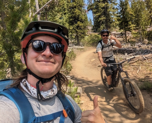 A male mountain biker gives a thumbs up to the camera with another mountain biker behind him